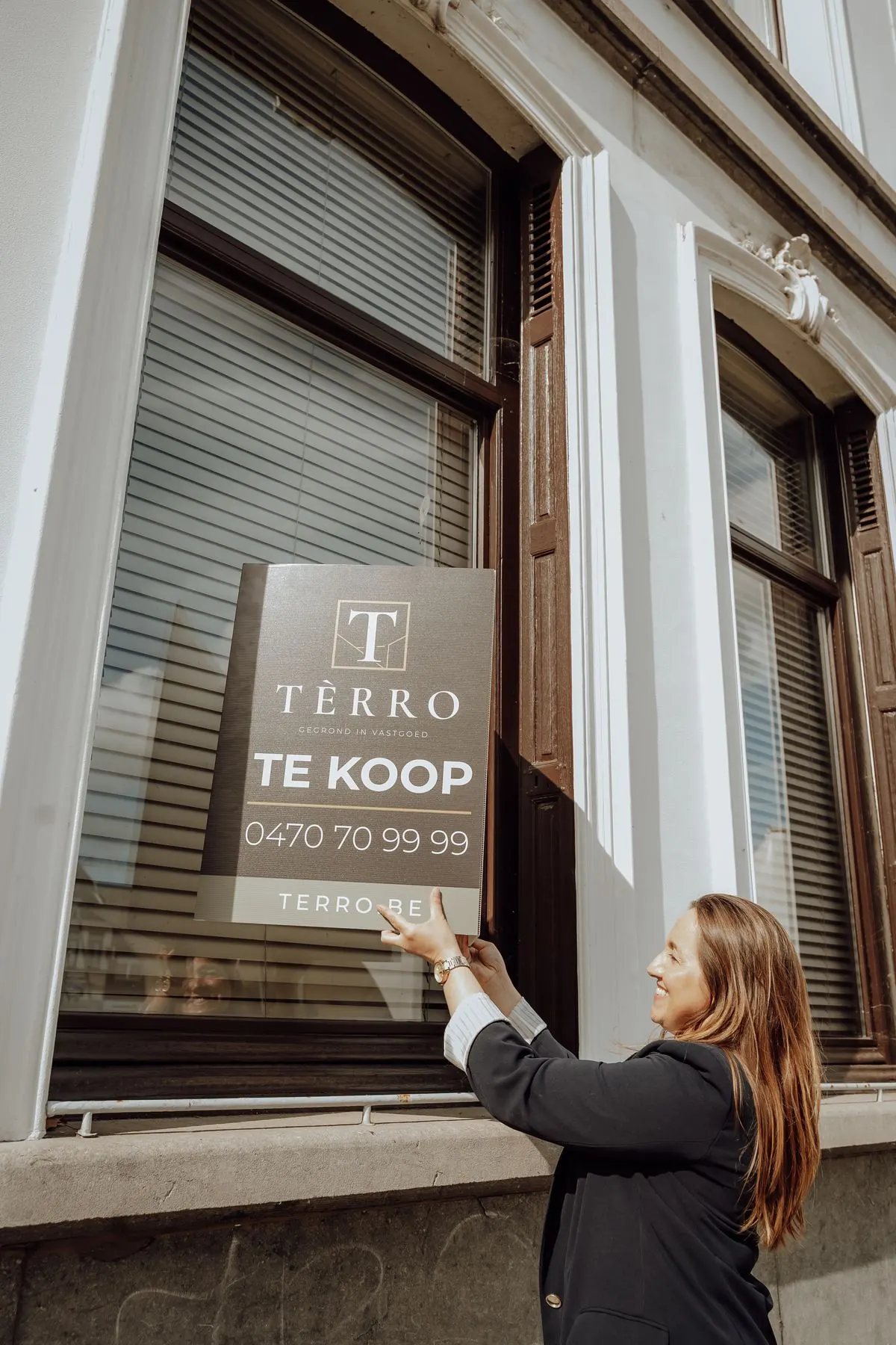 Makelaar van TÈRRO hangt een Te koop-bord aan het raam van een witte gevel